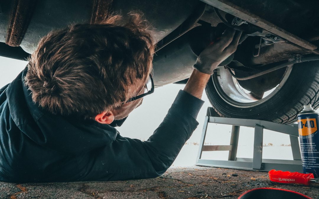 A person wearing glasses works diligently underneath a car, using tools in an auto shop. A tire and WD-40 can are visible nearby.