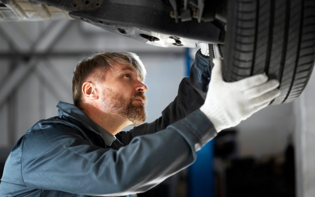 A mechanic in gloves inspecting a car tire and undercarriage in a garage.